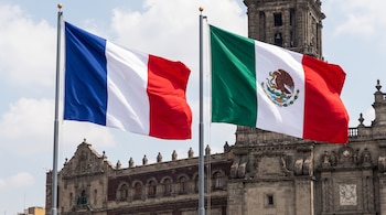 Las banderas de Francia y de México ondean en astas metálicas sobre el fondo de un edificio antiguo de piedra bajo un cielo claro.