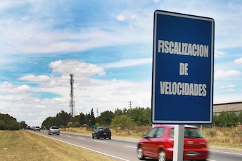 Un cartel azul con texto blanco 'FISCALIZACION DE VELOCIDADES' en primer plano, junto a un coche rojo en movimiento. Al fondo, una carretera con tráfico y una torre de telecomunicaciones bajo un cielo nublado