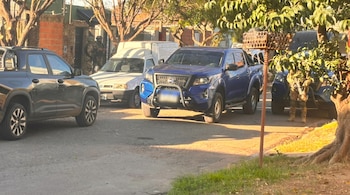 Vista de una calle de tierra con varias camionetas estacionadas, algunas azules, y un vehículo blanco. Personal uniformado se ve cerca de una casa de ladrillo.