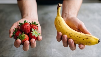 Dos manos sostienen fruta. Una mano sujeta cinco fresas rojas con tallos verdes. La otra mano sostiene un plátano amarillo maduro con manchas