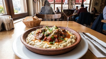 Un plato de Hünkârbeğendi con estofado de cordero, puré de berenjenas, queso y perejil, en una mesa de madera. Junto a él, una copa de vino, pan y cubiertos. Fondo borroso de restaurante.