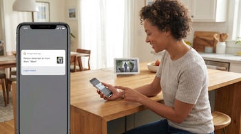 Mujer sonriente mirando su celular con la app Google Nest mostrando una alerta de persona detectada en la puerta. Un Google Nest Hub cercano también muestra la imagen.