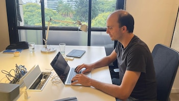 Guido Zack sentado en un escritorio blanco, tecleando en una laptop abierta. Detrás, una gran ventana muestra árboles verdes y edificios