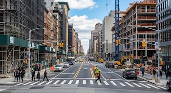 Vista de Flatbush Avenue en Brooklyn con edificios en construcción, andamios, tráfico de vehículos, peatones y una grúa bajo un cielo parcialmente nublado.