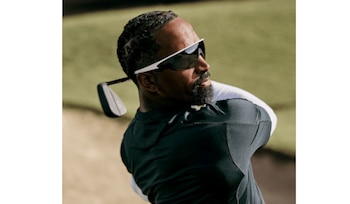 Hombre barbudo con gafas de sol blancas y oscuras, camiseta oscura, haciendo swing con un palo de golf en un campo con hierba y arena