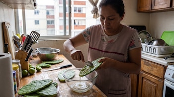 Una mujer con delantal extrae mucílago de un nopal con una cuchara. En la encimera de madera hay nopales, limas, un cuchillo y utensilios de cocina. Al fondo, una ventana y edificios.