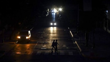 Vista nocturna de una calle oscura con dos personas cruzando un paso de cebra. Una furgoneta estacionada y varios vehículos avanzan con faros encendidos