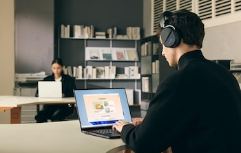 Un hombre de perfil con auriculares negros trabaja en una laptop oscura, sus manos en el teclado. Al fondo, una mujer usa una laptop blanca. Hay estanterías de libros