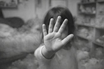 Primer plano en blanco y negro de la mano de un niño que cubre su rostro. Al fondo, un cuarto desordenado y borroso con estanterías y objetos.