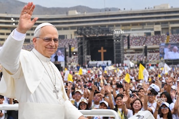 El Papa León XIV saluda con la mano levantada frente a una multitud en un estadio con un escenario y una cruz de madera al fondo