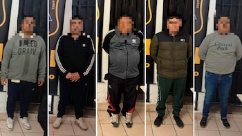 Cinco hombres, con rostros pixelados, de pie en fila frente a un fondo con un logo policial, vestidos con ropa casual
