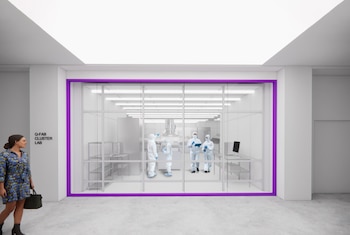 Interior de un laboratorio acristalado con marco morado donde personal con trajes de protección blanca trabaja cerca del rótulo Q-FAB CLUSTER LAB