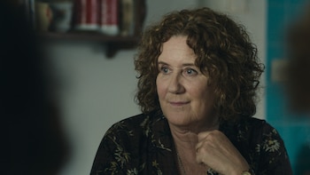 Una mujer de mediana edad con cabello rizado castaño y ojos azules claros, vestida con una camisa oscura estampada, mira hacia la izquierda