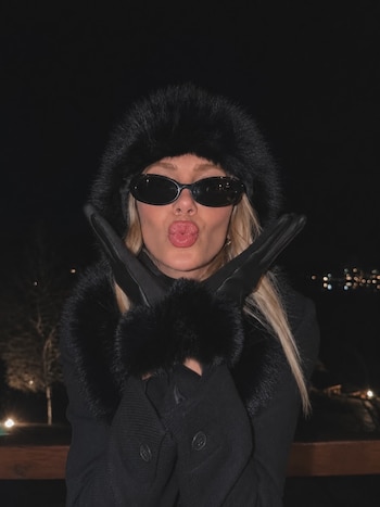 Una mujer rubia con abrigo negro, gorro de piel, gafas de sol y guantes cruzados frente al rostro posa de noche al aire libre
