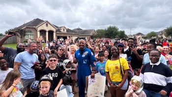 El astronauta Victor Glover, vestido con un traje de vuelo azul, sonríe y levanta el pulgar en medio de una multitud diversa que lo saluda con las manos en alto