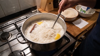 Una persona cocinando arroz con leche en una olla con cuchara sobre una cocina de gas. Incluye arroz seco en un tazón, envase de leche y nota escrita.