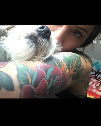 Una mujer con los brazos tatuados sostiene junto a su rostro a un perro pequeño de pelaje blanco