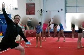 Un hombre con gafas y ropa deportiva negra posa junto a un grupo de niñas con rostros difuminados sobre una colchoneta roja en un gimnasio