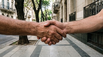Primer plano de dos manos de adultos estrechándose en un saludo, con una vereda de baldosas y edificios antiguos de Buenos Aires al fondo.