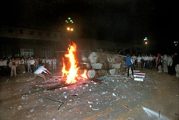 Un vehículo militar blindado arde intensamente en la oscuridad, rodeado por una multitud de personas. El suelo está cubierto de escombros y hay fuego brillante