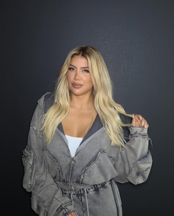 Wanda Nara posa de frente, con cabello rubio largo y suelto, vistiendo un enterito gris de tela lavada y un top blanco, frente a un fondo oscuro