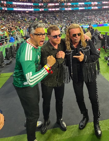 Tres hombres posan en el borde de un campo de fútbol. Detrás, un estadio lleno de público. Pantallas muestran "FIFA World Cup" y "Mexico City"