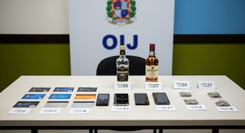 Mesa de madera clara con objetos decomisados: tarjetas bancarias, tres teléfonos, dos botellas de licor y paquetes de marihuana, todos etiquetados. Fondo borroso con logo OIJ.