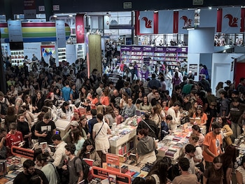 Vista aérea de una feria del libro con cientos de personas de todas las edades, navegando entre estantes y mesas llenas de libros, bajo luces y carteles