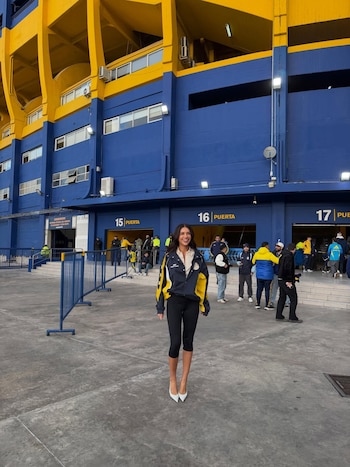 Mujer sonriente con chaqueta azul y amarilla, pantalones negros y tacones blancos, frente al estadio La Bombonera, con fachada azul y amarilla y gente en el fondo