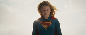 Retrato de Milly Alcock vestida como Supergirl, con un traje azul, capa roja y el escudo con la letra S. Cabello rubio y ondulado. Fondo de cielo claro