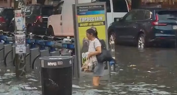 Mujer con camiseta a rayas y pantalones cortos camina por calle inundada. El agua le llega a las rodillas. Estación de bicicletas y varios vehículos están parcialmente sumergidos
