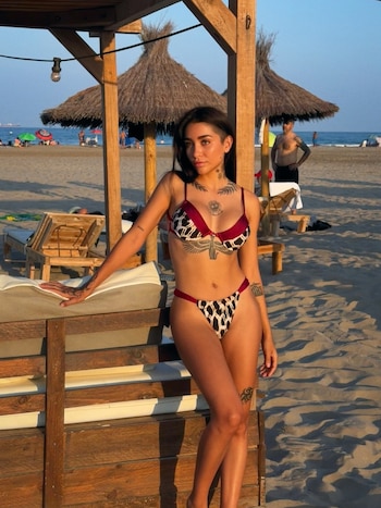 Mujer con bikini y tatuajes posa en una playa. Se ven sombrillas de paja, tumbonas de madera, mar, cielo azul y otras personas