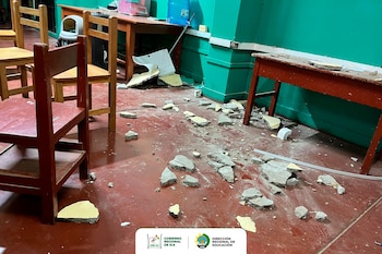 Un aula escolar con piso rojo, paredes verdes, sillas y mesas de madera. Hay escombros de concreto y yeso blanco esparcidos por todo el suelo, indicando daños estructurales