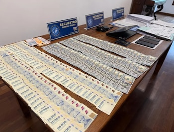 Pilas de billetes de 20.000 pesos, 100 dólares y euros, una laptop, smartphones, pasaporte, documentos y carteles de 'SECUESTRO DINERO' sobre una mesa de madera