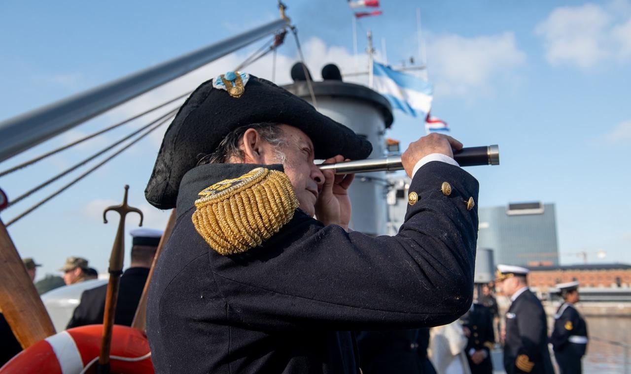 Un hombre vestido con un uniforme naval histórico y sombrero tricornio observa el horizonte a través de un catalejo en un barco, con la bandera argentina ondeando al fondo.