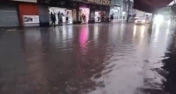 Vista a nivel de calle de una calle inundada en la capital, con agua cubriendo la vía frente a tiendas. Se ven personas en la acera y un coche con luces brillantes en la distancia
