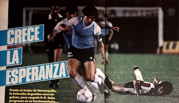 Un futbolista con camiseta celeste y blanca dribla el balón en un estadio, con un oponente caído y un árbitro cerca