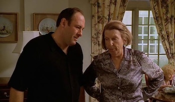 Un hombre calvo con camisa negra y una mujer mayor con blusa estampada están de pie en una habitación con cortinas florales y platos decorativos en la pared