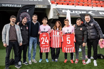 Un grupo de ocho personas, incluyendo los futbolistas Facundo Mura y Andrés Ayala con camisetas, posa en un campo de fútbol con gradas de estadio al fondo