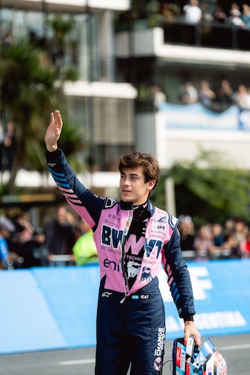 Hombre joven con mono de carreras rosa y azul oscuro, saluda con la mano derecha levantada mientras sostiene un casco. Fondo borroso de público y edificios