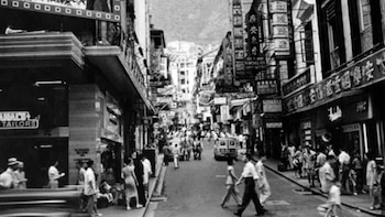 Foto en blanco y negro de una calle urbana con edificios altos y tiendas, llena de peatones en aceras y calzada, con un vehículo en el fondo