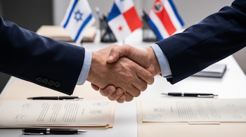 Plano cerrado de dos personas en traje oscuro dándose la mano sobre documentos oficiales y bolígrafos. Al fondo, banderas desenfocadas de Israel, Panamá y Costa Rica.