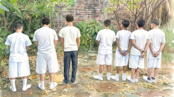 Pintura de acuarela con ocho personas de espaldas; siete niños de blanco y un adulto de pantalón oscuro, contra pared de ladrillo y vegetación.