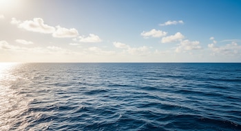 Océano con aguas azules, pequeñas olas, cielo claro y nubes blancas dispersas. La luz del sol se refleja sobre el agua.