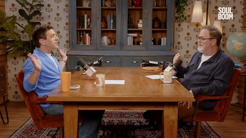 B.J. Novak y Rainn Wilson sentados frente a frente en una mesa de madera con micrófonos, conversando en un estudio con estantería y papel tapiz