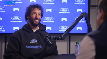 Daniel Ricciardo sonriendo, con cabello rizado y barba, usando una sudadera negra con logos de Ford Racing, sentado frente a un micrófono