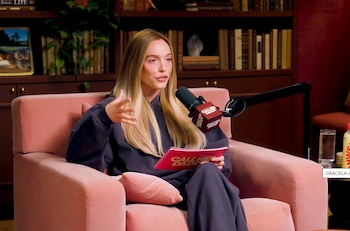 Mujer rubia sentada en un sillón rosa, hablando por un micrófono con el logo «Call Her Daddy» y sosteniendo un cuaderno rosa del podcast, con estanterías al fondo