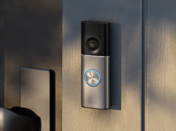 Un Ring Video Doorbell Wired gris y negro con un botón circular azul brillante está montado verticalmente en una pared gris claro texturizada, junto a una puerta oscura