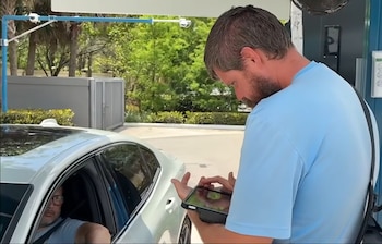 Un hombre barbudo en una camiseta azul claro opera una tableta mientras interactúa con un cliente en un coche compacto blanco en un lavadero de autos al aire libre