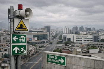 Vista aérea de una ciudad peruana bajo cielo nublado. Primer plano: poste con sirena roja y señales de riesgo sísmico y evacuación. Fondo: edificios, calles y universidad.